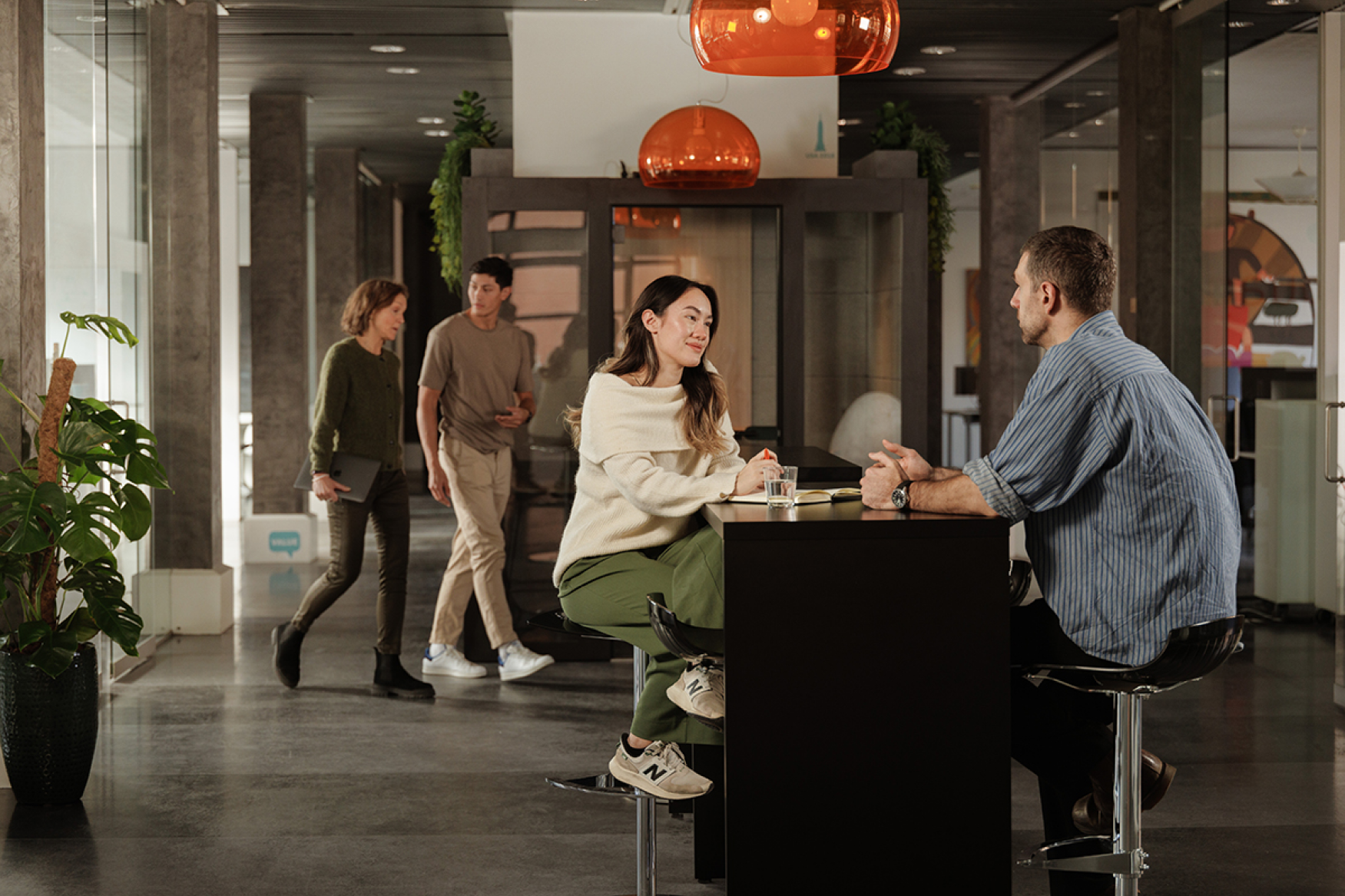 Colleagues sitting at a high table in an office having a discussion, while two others walk by in the background.