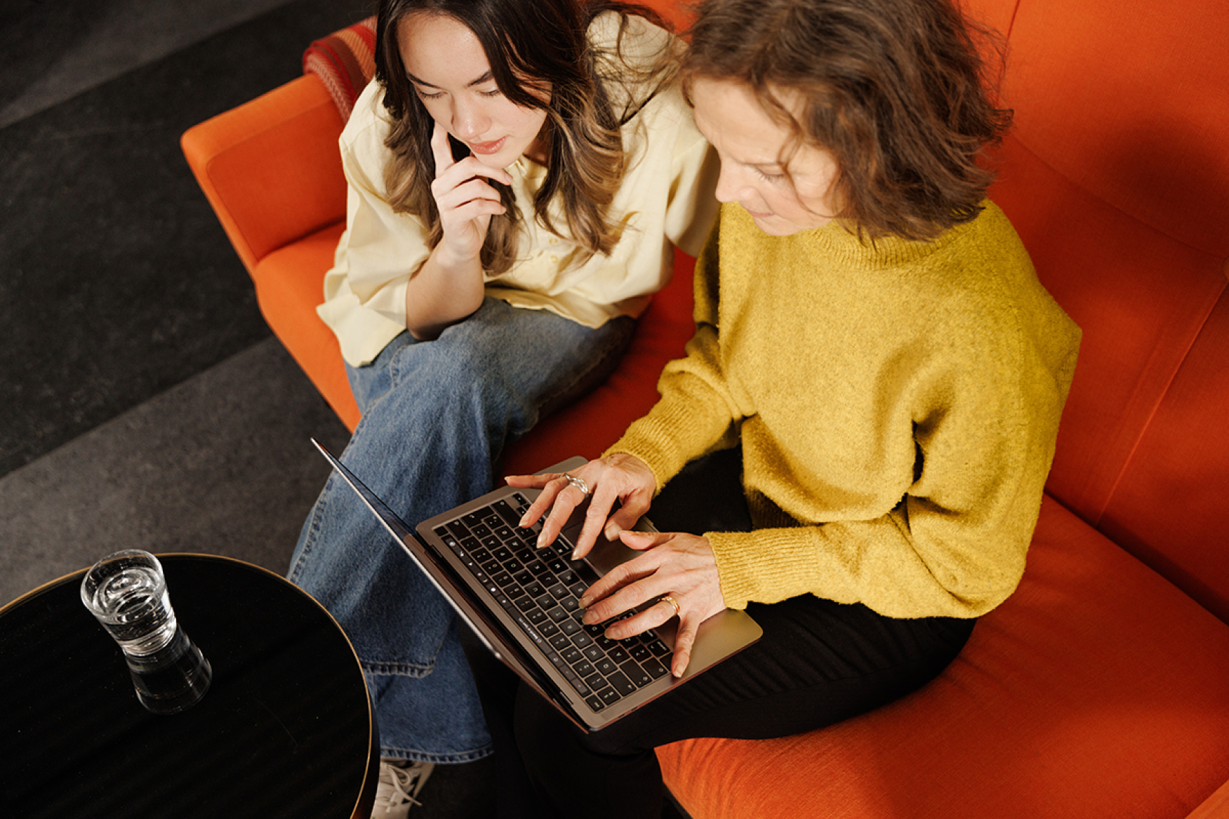 Two women sitting on an orange couch looking intently at a laptop