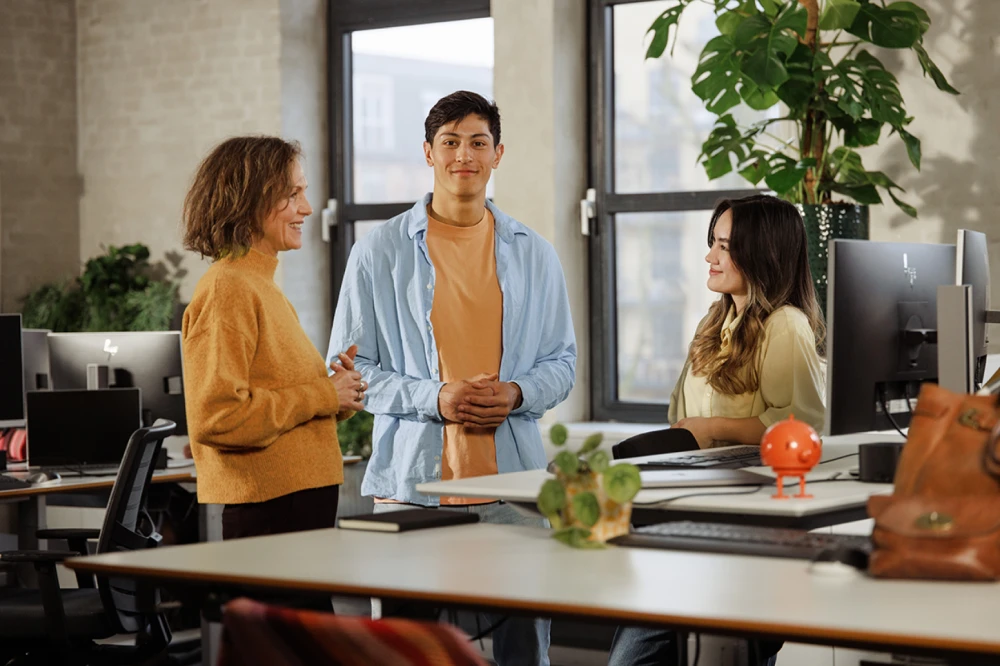 Two women talking to each other while a male colleague stands between them, looking at the camera.