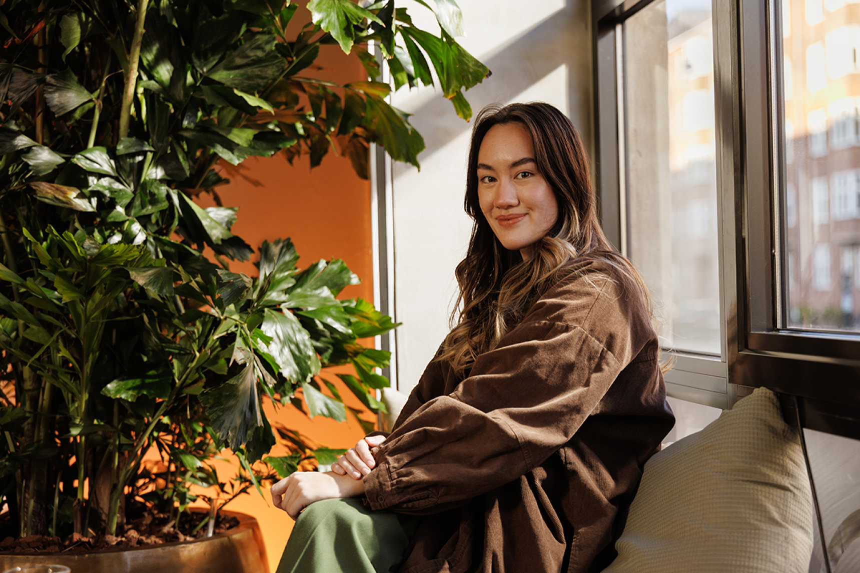 Woman sitting and smiling at the camera, surrounded by a large office plant and warm orange lighting.