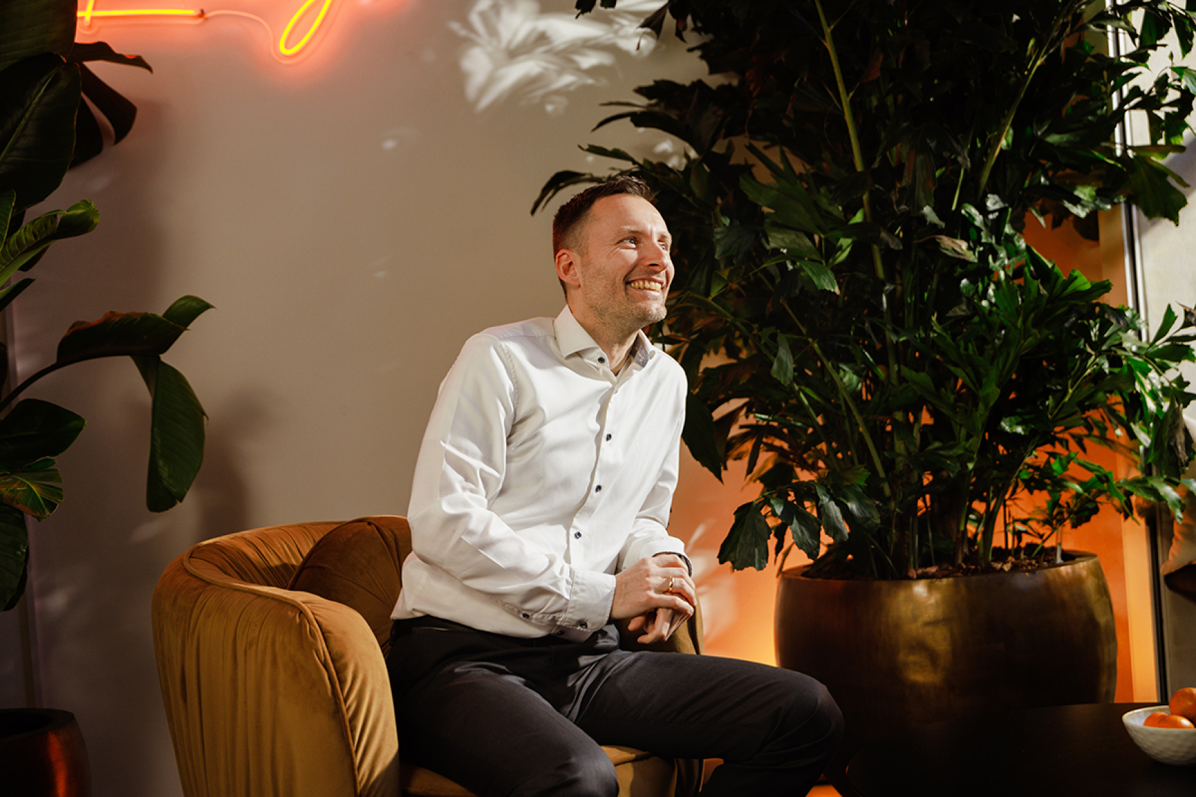 Man sitting in a chair smiling with office plants and warm orange lighting in the background