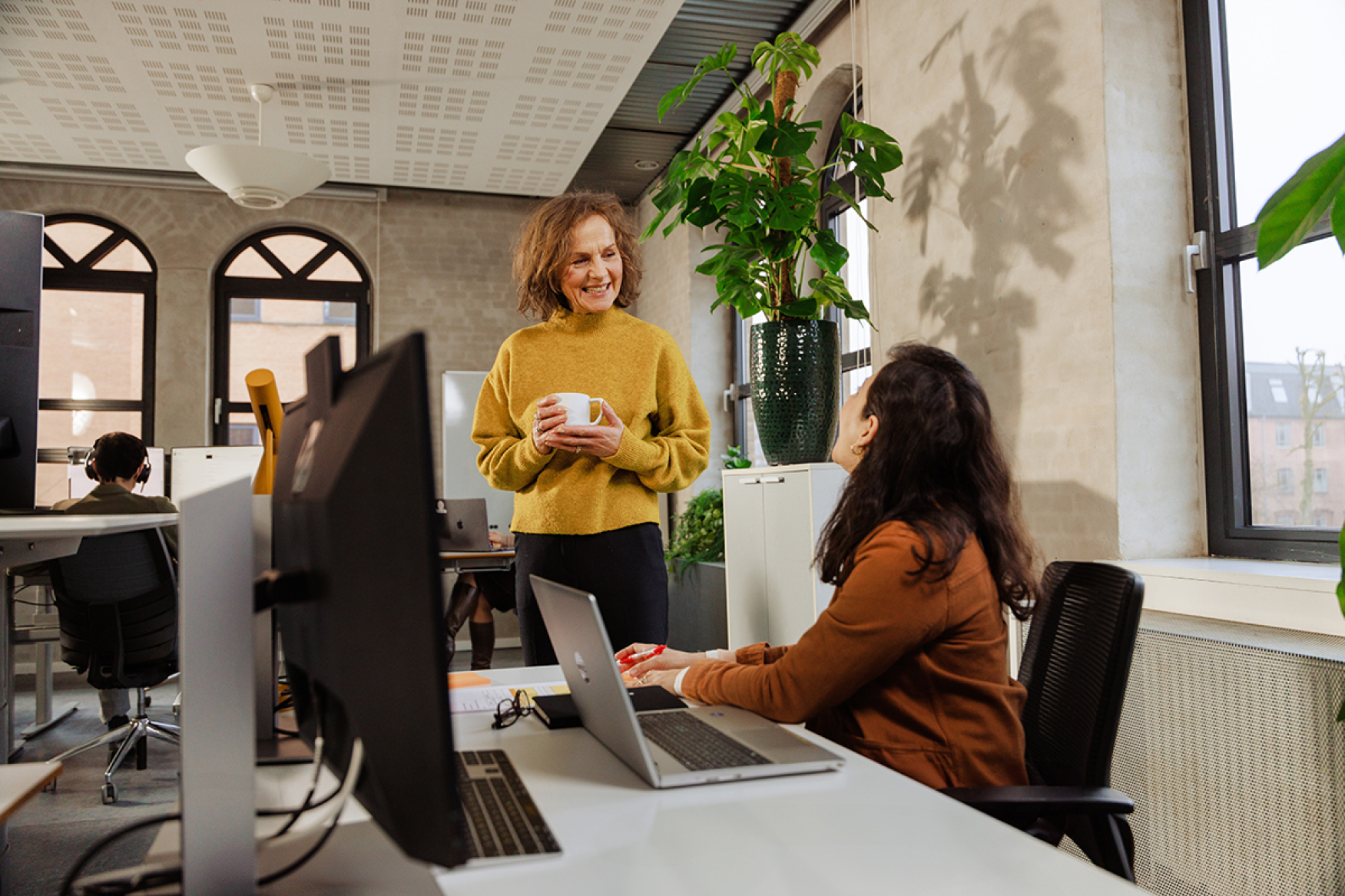Two female coworkers talking at their desks, one standing and holding a coffee cup.