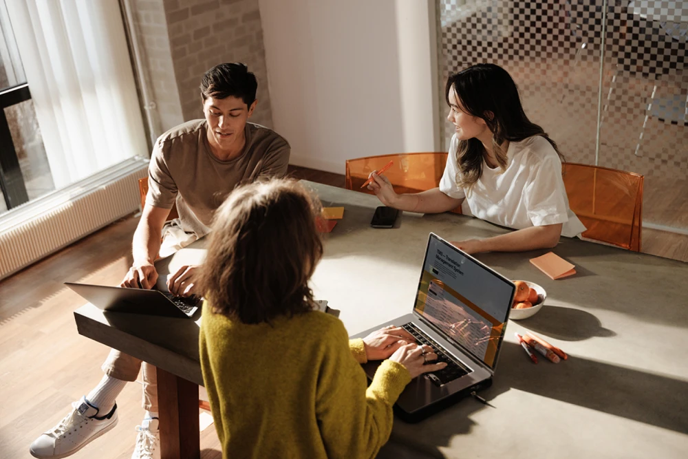 Three coworkers in a meeting talking with their laptops while sitting at a table