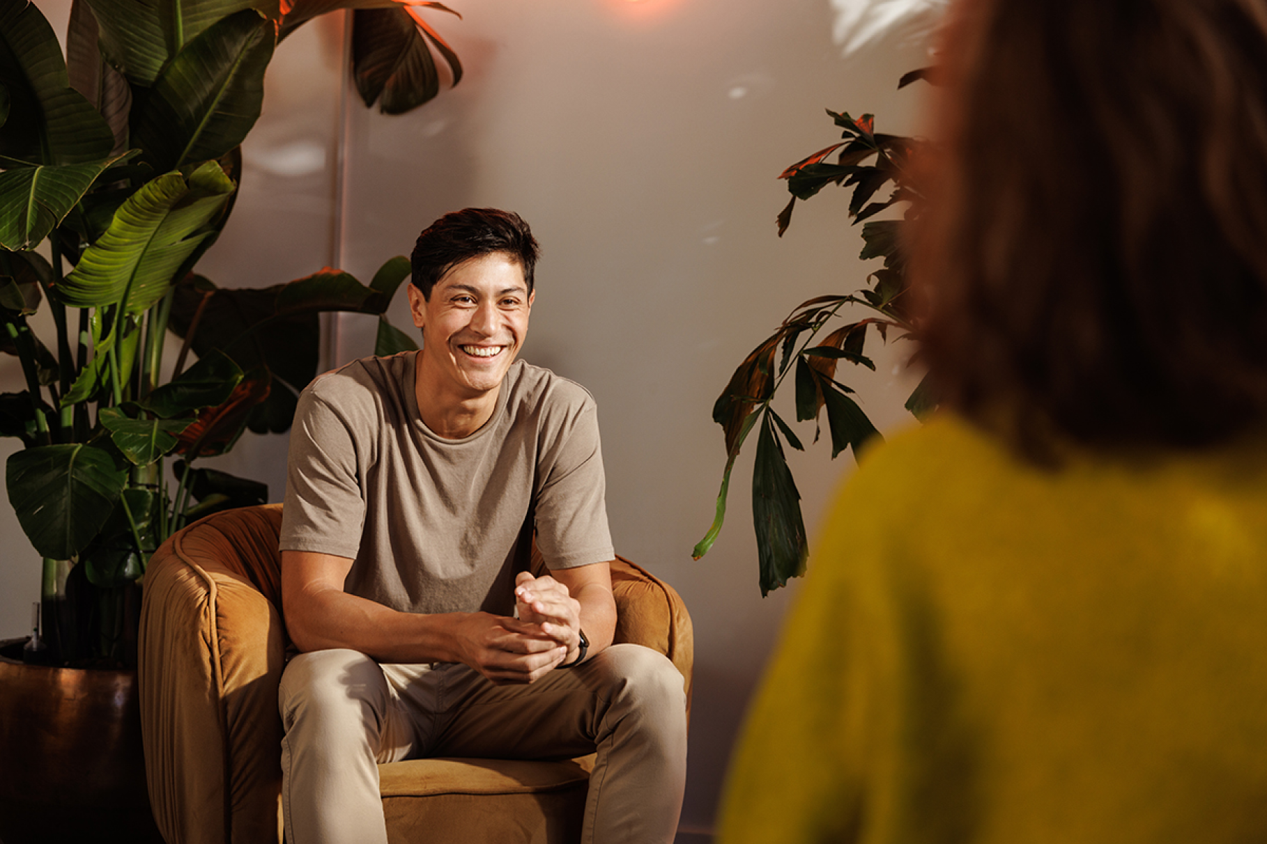 Young man sitting in chair surrounded by office plants smiling in conversation