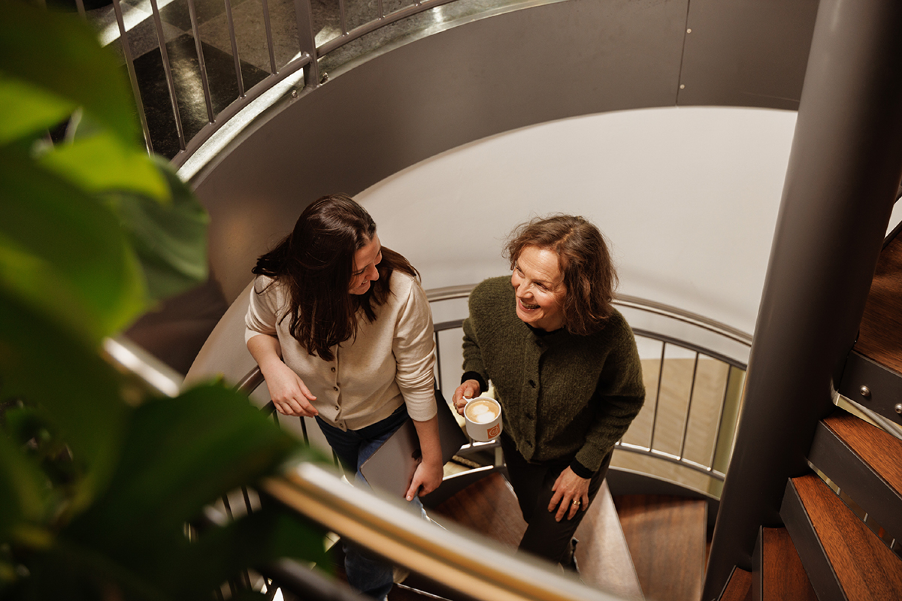 Two women walking up a set of spiral stairs having a discussion while holding coffee and a laptop